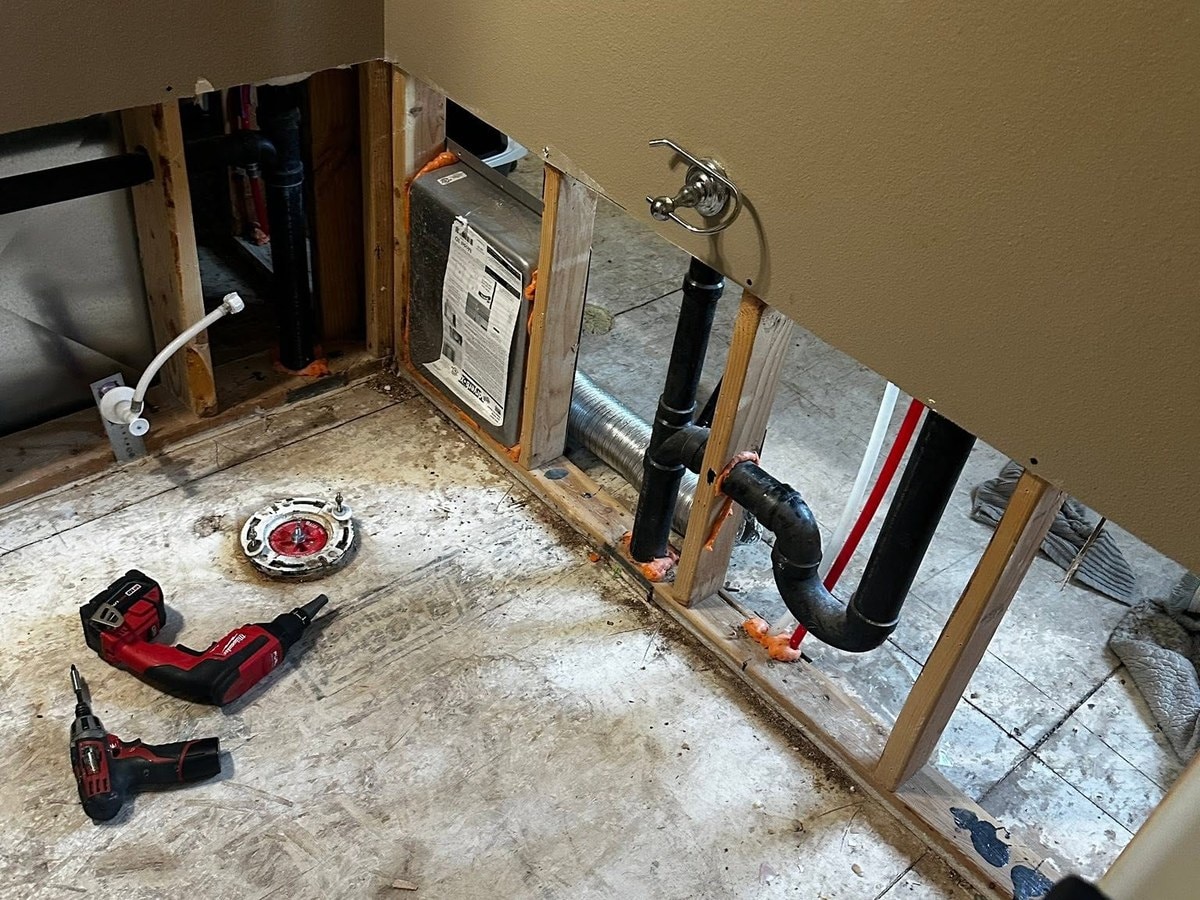 Bathroom stripped to bare subfloor with the wall cut open at the base and the plumbing exposed