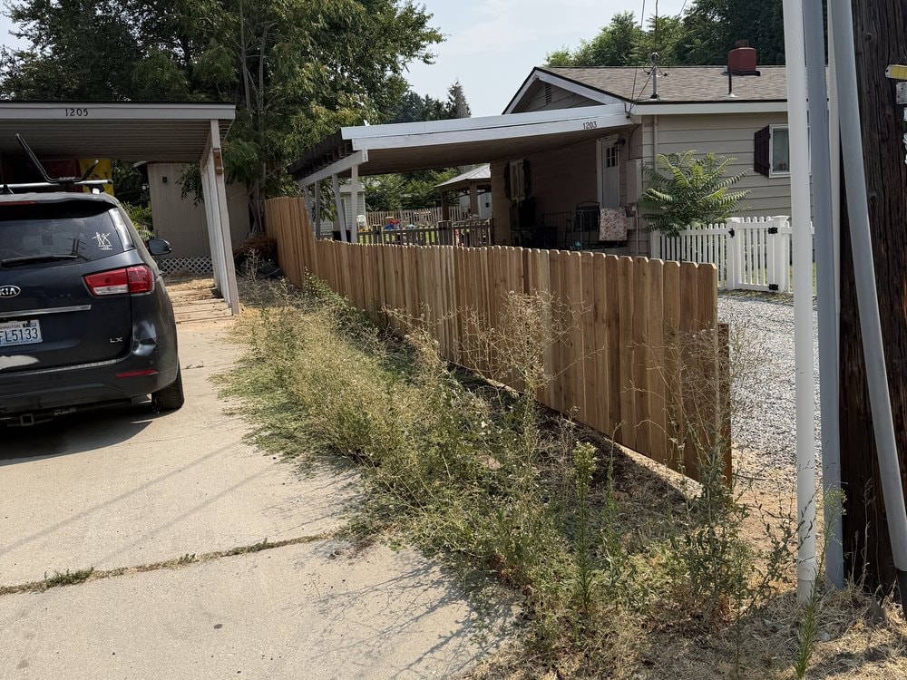 Light cedar fence running past the carport with dry weeds still standing at its base