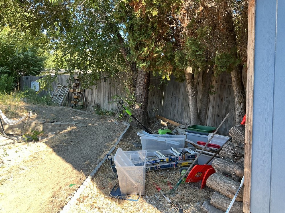 Weathered gray privacy fence behind stacked storage totes, a ladder, hand tools and cut logs