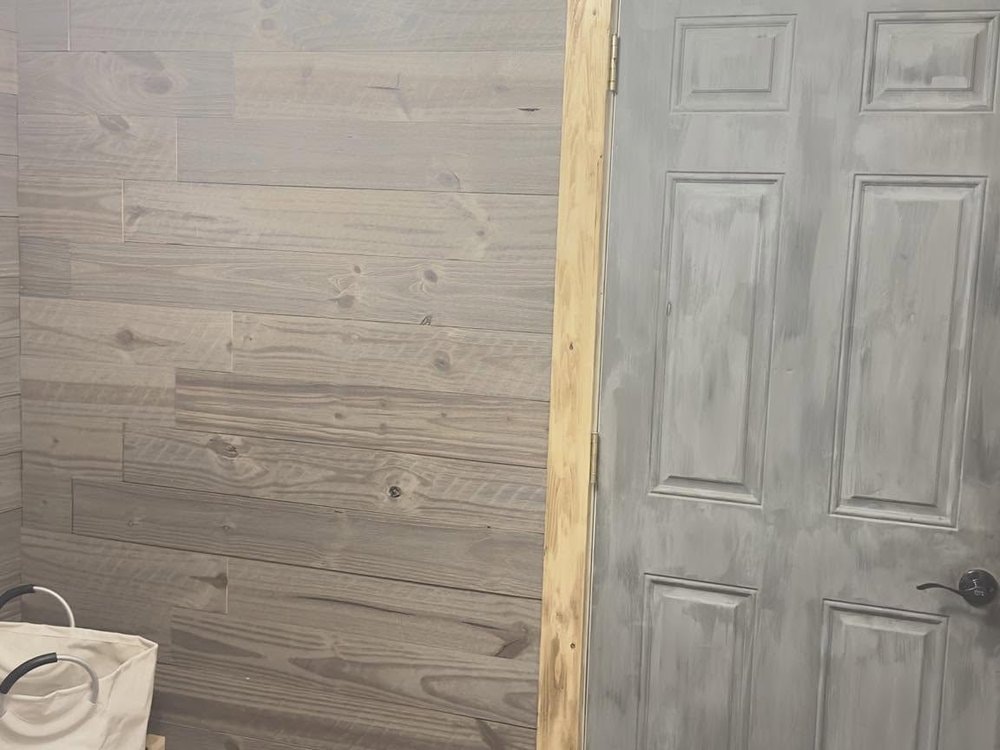 Gray plank wall meeting a gray six-panel door in raw wood trim, laundry bag and white appliance at left