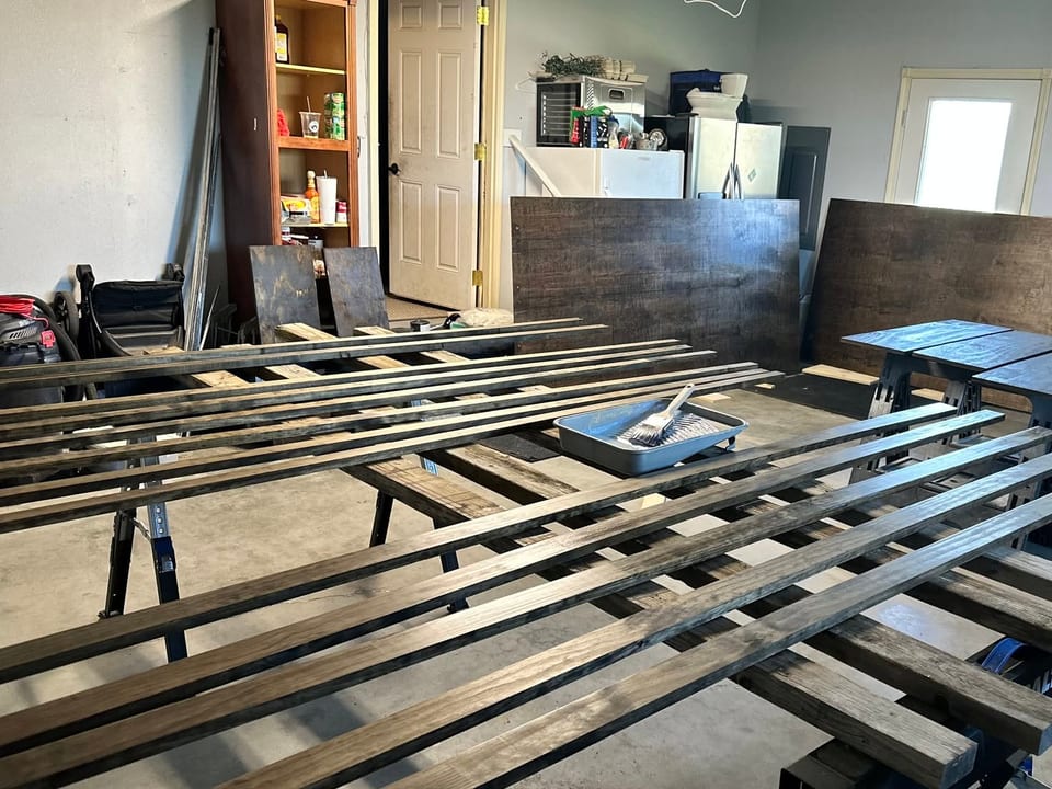 Long stained boards on sawhorses in a garage with a stain tray, brush, and dark panels.