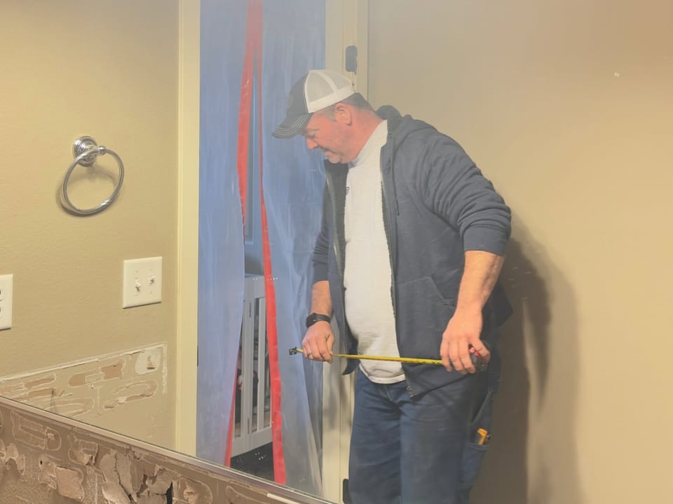 Plastic sheeting hangs in the doorway as a worker measures a wall stripped to torn paper and adhesive.