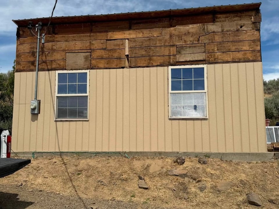 Tan vertical panels cover the lower wall of an outbuilding, with weathered wood boards still exposed above.