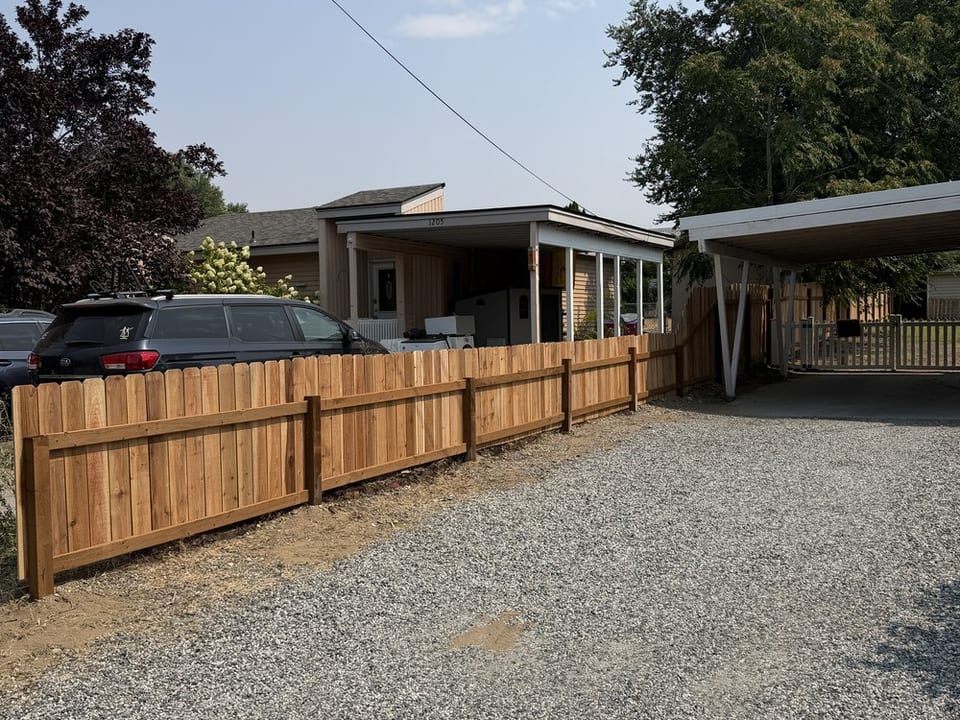 Freshly built wood privacy fencing with square posts runs alongside a gravel driveway past a house toward a carport.