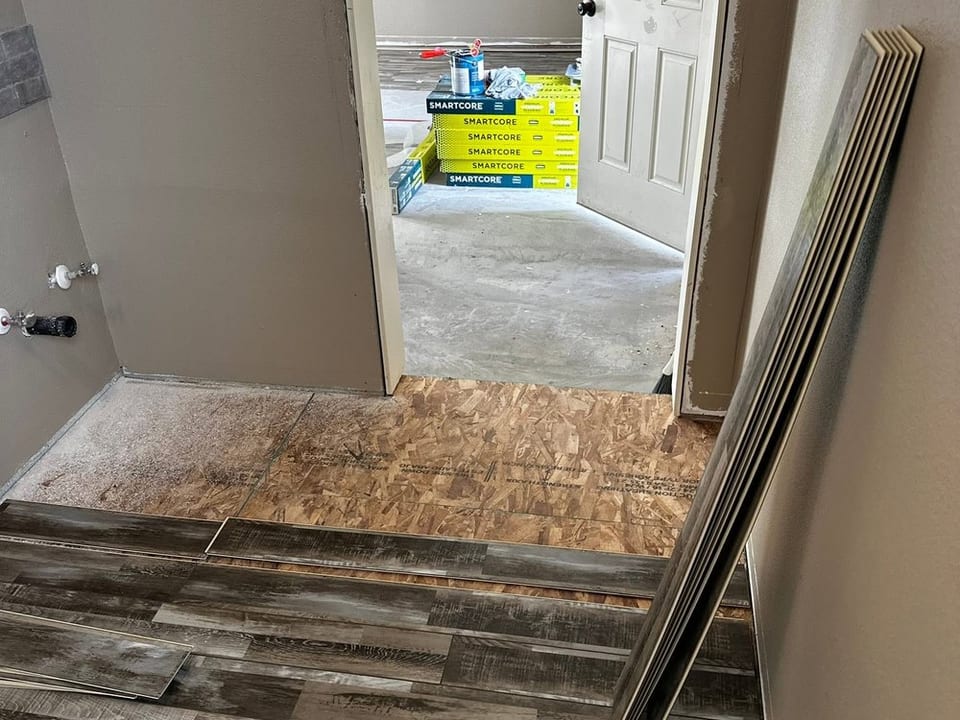 Grey vinyl planks partly laid up to a doorway, bare subfloor exposed, spare planks leaning against the wall.
